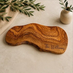 May include: An irregular, organic-shaped olive wood serving board featuring a natural live edge and prominent wood grain patterns. The board is engraved with the text The Williams in a cursive script font on the lower right side of the surface.