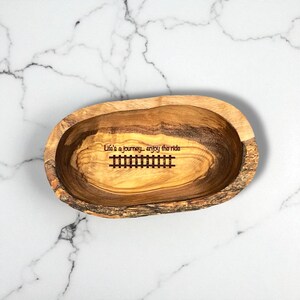 May include: An oval-shaped wooden bowl with a natural bark edge and visible wood grain. The interior features the engraved text "Life's a journey... enjoy the ride" positioned above a graphic illustration of a set of railroad tracks.
