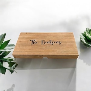 K&ouml;nnte beinhalten: Eine rechteckige, hellbraune Holzschachtel mit glatter Maserung. Auf der Oberseite ist der Text "The Bentsens" in einer schwarzen Schreibschrift eingraviert. Die Schachtel hat ein schlichtes, flaches Design mit einer kleinen Kerbe an der Vorderkante.