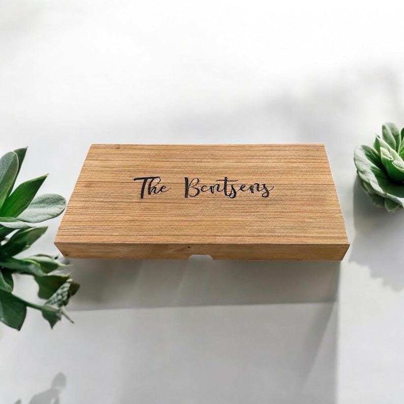 K&ouml;nnte beinhalten: Eine rechteckige, hellbraune Holzschachtel mit glatter Maserung. Auf der Oberseite ist der Text "The Bentsens" in einer schwarzen Schreibschrift eingraviert. Die Schachtel hat ein schlichtes, flaches Design mit einer kleinen Kerbe an der Vorderkante.