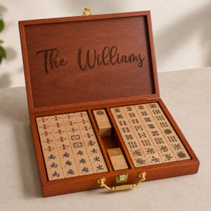 K&ouml;nnte beinhalten: Ein Mahjong-Spiel aus Holz in einem rechteckigen Koffer mit goldenem Verschluss und Griff. Auf der Innenseite des Deckels ist der Text The Williams eingraviert. Das Set enth&auml;lt zahlreiche Spielsteine mit blauen und roten chinesischen Schriftzeichen.