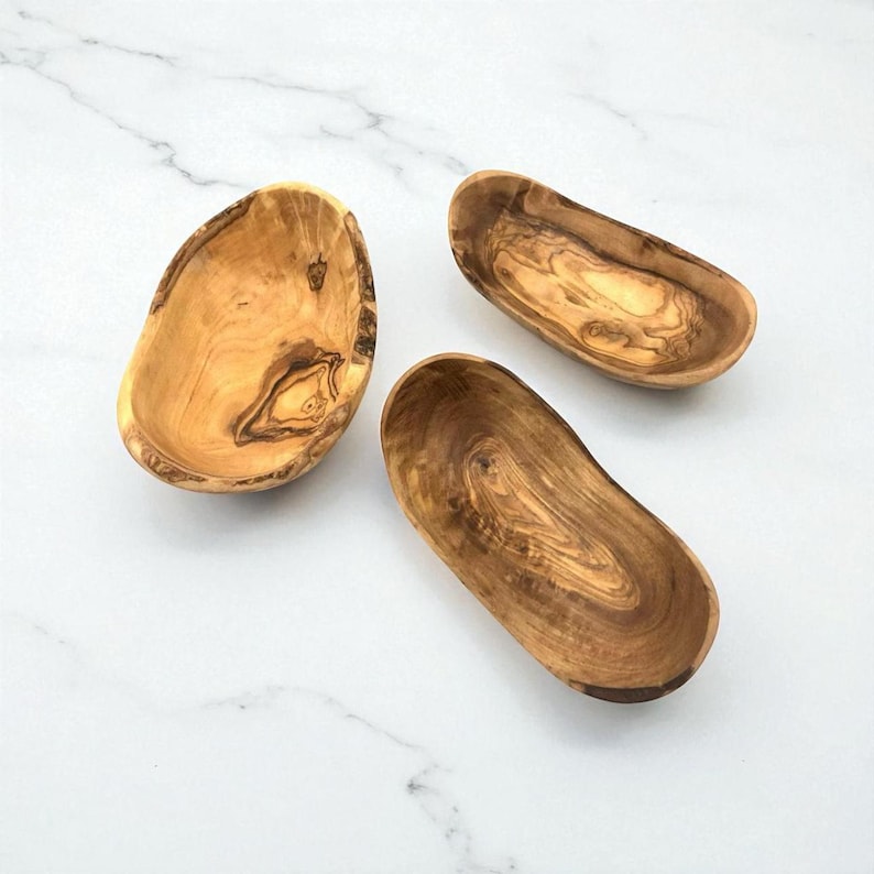 May include: Three small, shallow bowls crafted from light brown olive wood with prominent dark brown grain patterns. The bowls feature irregular, organic shapes with smooth, curved edges and a polished finish, showcasing the natural texture of the wood.