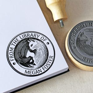 Op de afbeelding: Een zwart-witte stempel met een kattenontwerp en de tekst "FROM THE LIBRARY OF MEGAN FOSTER". De stempel ligt op een wit oppervlak naast een houten handvat en een afdruk.