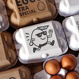 May include: An overhead shot of several egg cartons. One carton is white with a cartoon egg wearing sunglasses and the word "FRESH". Other cartons are brown with the words "EGG EXTRA SPECIAL". Several brown eggs are visible.