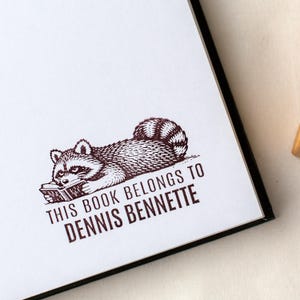 May include: A white book page featuring a brown illustration of a raccoon reading a book. The text "THIS BOOK BELONGS TO DENNIS BENNETTE" is printed below the raccoon image.