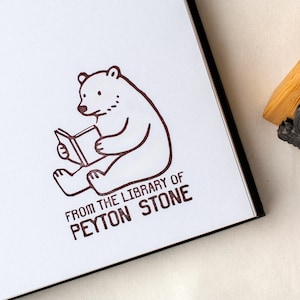 Puede incluir: Una página blanca con una ilustración en tinta marrón de un oso leyendo un libro. El texto "FROM THE LIBRARY OF PEYTON STONE" está impreso debajo del oso. La imagen está sobre una superficie blanca.