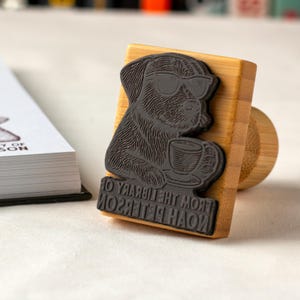May include: A wooden stamp featuring a dog wearing sunglasses and holding a coffee cup. The stamp has the text "FROM THE LIBRARY OF NOAH PETERSON" in a curved font. The stamp is made of wood and dark gray rubber.