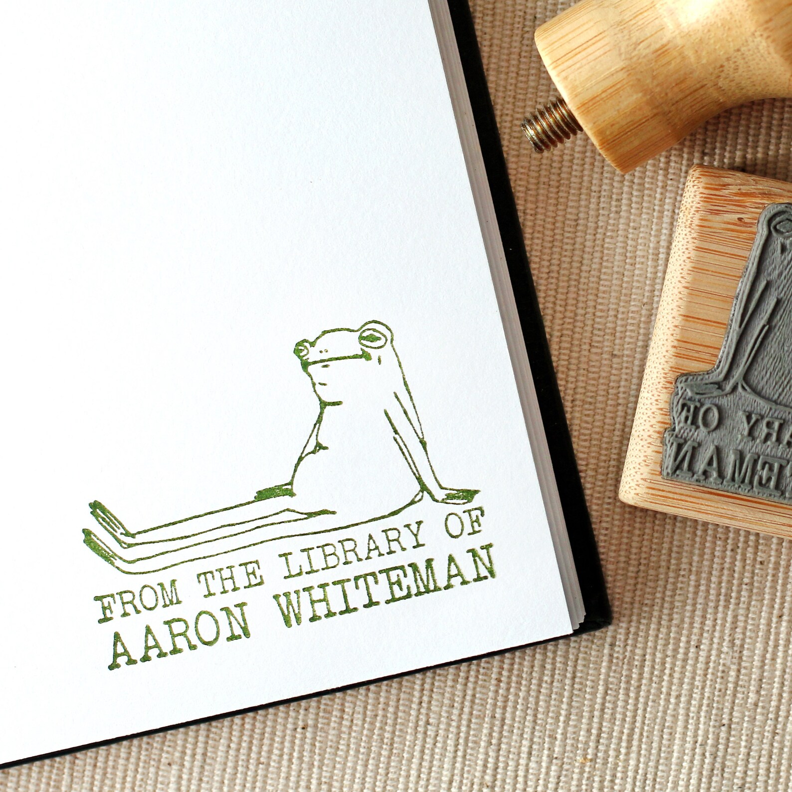 A close up of the bottom right page of a book, with a green stamp of a frog sitting like a human that says "From the library of Aaron Whiteman" underneath.