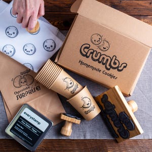 May include: A collection of branding supplies including a cardboard box labelled Crumbs Homemade Cookies, brown paper bags with Chocolate Surprise text, paper cups with Crumbs branding, a black ink pad, and wooden stamps with custom designs.