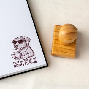 May include: A wooden stamp with a bamboo handle next to a notebook with a dog illustration. The stamp features a dog wearing sunglasses and holding a coffee cup, with the text "FROM THE LIBRARY OF NOAH PETERSON" below the image.