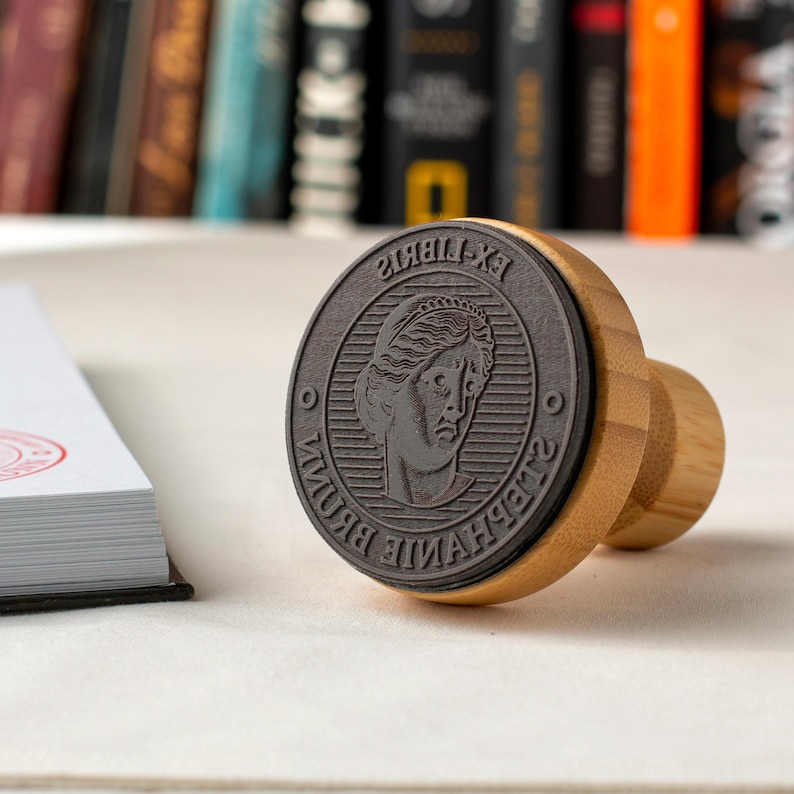 May include: A circular, dark grey stamp featuring a classical female bust and the text "EX LIBRIS STEPHANIE BROWN". The stamp has a natural wood handle. A stack of white paper is in the background.