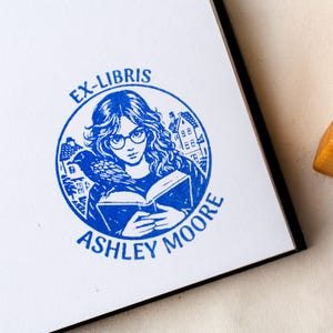 May include: A blue and white Ex-Libris stamp featuring a woman with glasses reading a book, a raven, and houses. The stamp includes the text "EX-LIBRIS" at the top and "ASHLEY MOORE" at the bottom, all in a circular design.