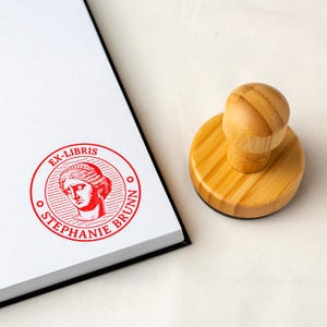 May include: A custom stamp in red and white, featuring the text "EX-LIBRIS STEPHANIE BRUNN" around a classical bust. The stamp has a round wooden handle and is beside a stack of white paper.