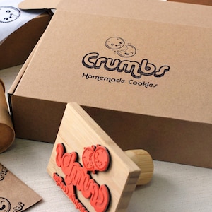 May include: A brown cardboard box with the text Crumbs Homemade Cookies and a cookie graphic, a matching paper cup, a wooden rubber stamp with red text, and branded tissue paper featuring small cookie illustrations and the Crumbs logo.