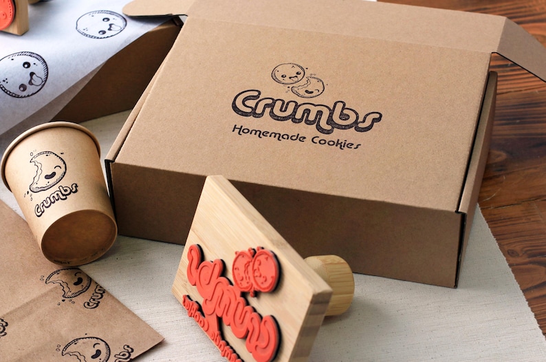 May include: A brown cardboard box with the text Crumbs Homemade Cookies and a cookie graphic, a matching paper cup, a wooden rubber stamp with red text, and branded tissue paper featuring small cookie illustrations and the Crumbs logo.