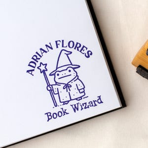 Könnte beinhalten: Ein blauer Tintenstempel auf weißem Papier zeigt einen Cartoon-Frosch-Zauberer mit einem sternförmigen Zauberstab. Der Text "ADRIAN FLORES" wölbt sich über dem Frosch, darunter "Book Wizard". Der Stempel befindet sich auf einer weißen Oberfläche.
