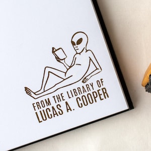 May include: A brown ink stamp on white paper features an alien reading a book. The text "FROM THE LIBRARY OF LUCAS A. COOPER" is printed below the alien illustration. The stamp is on a book or journal.