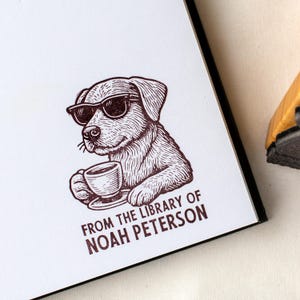 May include: A brown ink stamp featuring a dog wearing sunglasses and holding a teacup. The text "FROM THE LIBRARY OF NOAH PETERSON" is printed below the dog. The stamp is on a white surface.