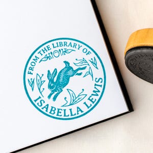 Può includere: Un timbro circolare turchese con la scritta "FROM THE LIBRARY OF ISABELLA LEWIS" che circonda un'illustrazione di un coniglio che salta. Il timbro è su carta bianca, con un oggetto giallo e nero sullo sfondo.
