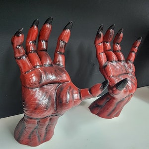 Könnte beinhalten: Zwei rote, gekrallte Hände mit schwarzen Fingernägeln, in einer bedrohlichen Pose modelliert. Die Hände sind mit strukturierter Haut detailliert und scheinen aus einem strapazierfähigen Material zu sein.
