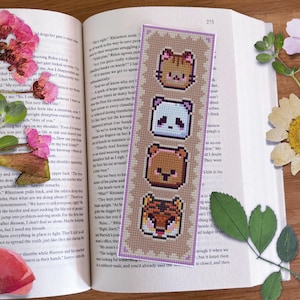 May include: A purple and beige bookmark with a cross-stitch design featuring four animal faces: a cat, a panda, a bear, and a tiger. The bookmark is on a book with text.