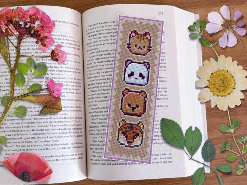 May include: A purple and beige bookmark with a cross-stitch design featuring four animal faces: a cat, a panda, a bear, and a tiger. The bookmark is on a book with text.