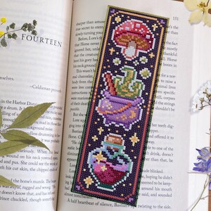 May include: A purple, green, and red cross-stitch bookmark with a cauldron, a potion bottle, and a mushroom. The background is a dark purple with stars.