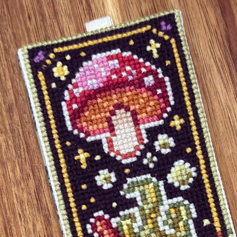 May include: A cross-stitch bookmark with a mushroom design. The mushroom is red and white with a brown stem. The background is black with gold stars and white flowers.