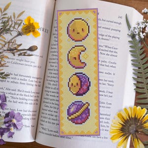 May include: A yellow bookmark with a cross-stitch design of celestial bodies. The bookmark features a yellow sun, a crescent moon, a half moon with a purple crescent, and a purple planet with a gold ring.