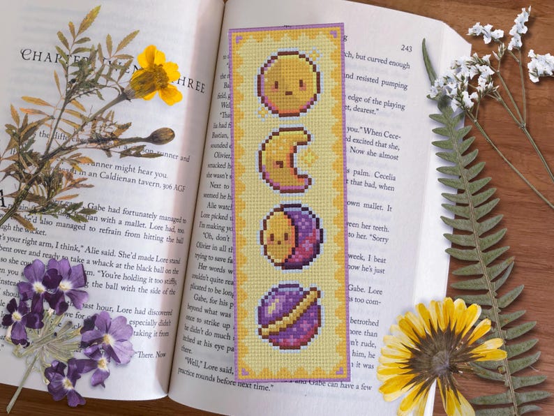 May include: A yellow bookmark with a cross-stitch design of celestial bodies. The bookmark features a yellow sun, a crescent moon, a half moon with a purple crescent, and a purple planet with a gold ring.