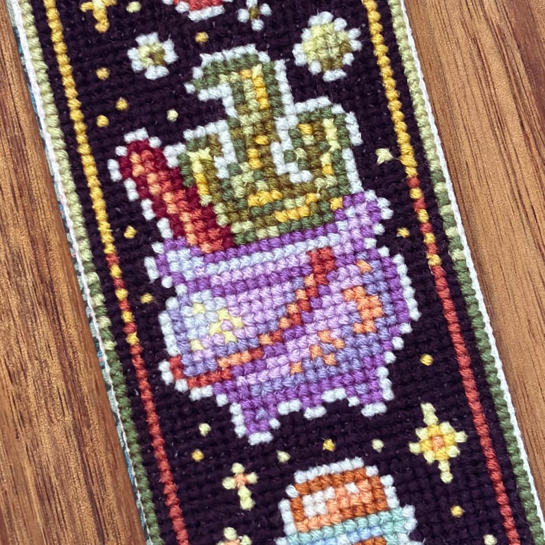 May include: A cross-stitch pattern of a purple pot with a green cactus plant inside. The pot is decorated with small yellow stars. The background is black with yellow stars.