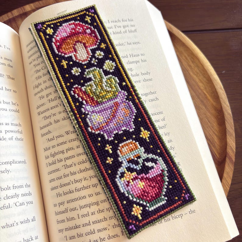 May include: A cross-stitch bookmark with a dark purple background and a green border. The bookmark features a mushroom, a cauldron, and a potion bottle with a pink and red liquid. The design is surrounded by stars and other small details.