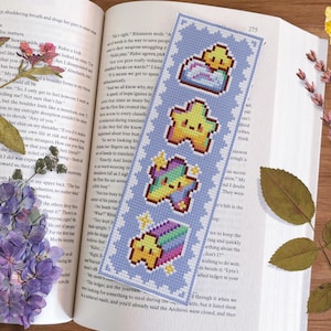 Puede incluir: Un marcapáginas de punto de cruz azul y blanco con un diseño caprichoso que presenta cuatro estrellas con diferentes colores y patrones. El marcapáginas está decorado con un borde blanco y una pequeña estrella blanca en la esquina superior derecha.