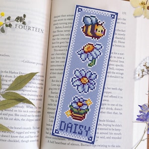 Peut inclure: Un marque-page en point de croix bleu et blanc avec un motif de marguerite et le mot "DAISY" au centre. Le marque-page présente également une abeille, un pot de fleurs et une fleur.