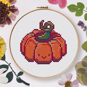 Op de afbeelding: Een kruissteekontwerp van een pompoen in een borduurring. De pompoen is oranje met een groene stengel en een lachend gezicht. De achtergrond is wit en de ring is goudkleurig. Gedroogde bloemen omringen de ring.