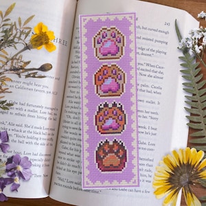 May include: A purple and white bookmark with a cross-stitch design of four paw prints. The paw prints are brown, pink, and white.
