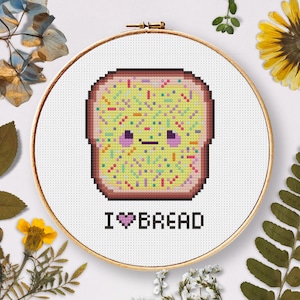 Puede incluir: Un aro de bordado blanco con un diseño de punto de cruz de una rebanada de pan de dibujos animados con chispas y una cara sonriente. El texto "I ♥ BREAD" está debajo del pan.