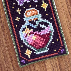 May include: A close-up of a colorful embroidered potion bottle with a green, white, and orange stopper. The bottle is filled with a pink and red liquid and surrounded by yellow stars on a black background. The design is framed with a green, orange, and brown border.