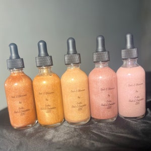 Puede incluir: Cinco botellas de vidrio con goteros llenas de un líquido brillante. Las botellas están etiquetadas "Bad & Shimmer by Erika Bad-E Cosmetics 220".