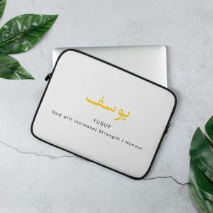 May include: White laptop sleeve with black edging, displaying the name "YUSUF" in gold Arabic script. The English translation below reads "God will increase Strength | Honour". The sleeve is on a light grey surface with green leaves.