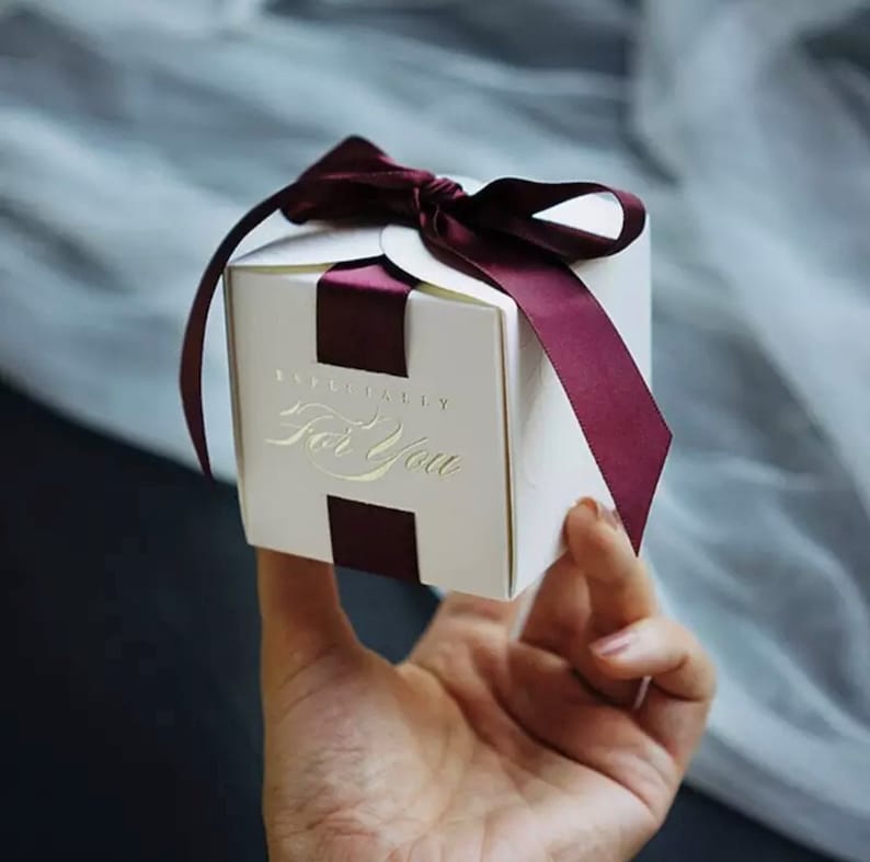 May include: A small, white gift box with a burgundy ribbon and the words "Especially For You" in gold lettering. The box is held in a person's hand, with the ribbon tied in a bow.