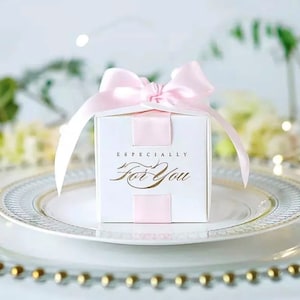 May include: A white square gift box with a pink satin bow and ribbon. The box has the words "Especially For You" in gold script. The box is sitting on a white plate with a gold rim, suggesting a celebratory occasion.