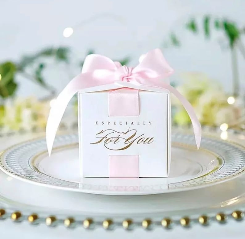 May include: A white square gift box with a pink satin bow and ribbon. The box has the words "Especially For You" in gold script. The box is sitting on a white plate with a gold rim, suggesting a celebratory occasion.