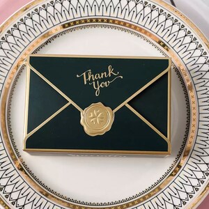 May include: A green envelope-shaped box with a gold border and a gold wax seal with a four-leaf clover design. The box has the words "Thank You" written in gold script on the front.