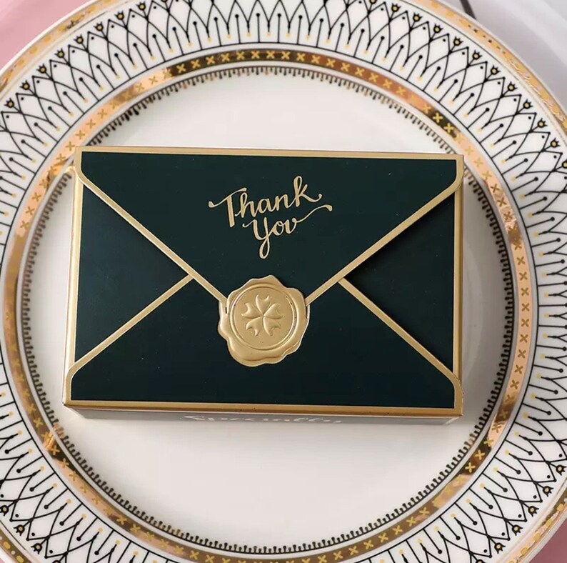 May include: A green envelope-shaped box with a gold border and a gold wax seal with a four-leaf clover design. The box has the words "Thank You" written in gold script on the front.