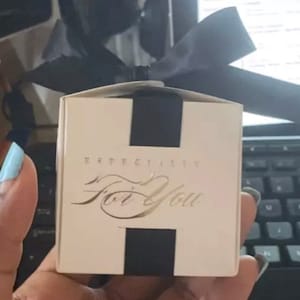 May include: A small, square gift box with a black ribbon and accents. The box is cream-colored with the words "Especially For You" in gold script. The box is held in a hand.