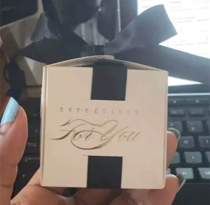 May include: A small, square gift box with a black ribbon and accents. The box is cream-colored with the words "Especially For You" in gold script. The box is held in a hand.
