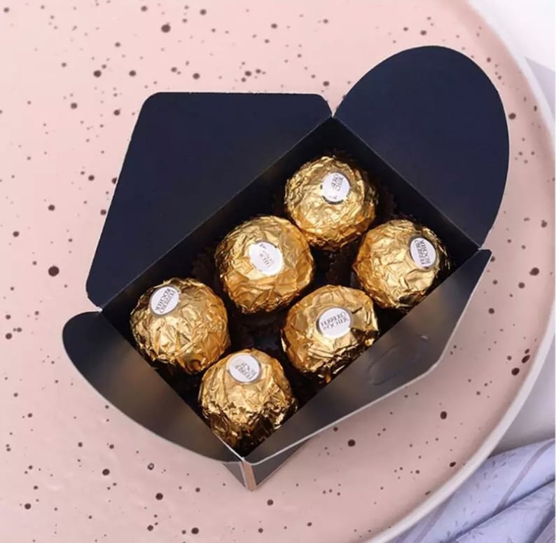 May include: A black gift box with a flap top, filled with six gold-wrapped chocolate candies. The candies have the words "Ferrero Rocher" printed on them.