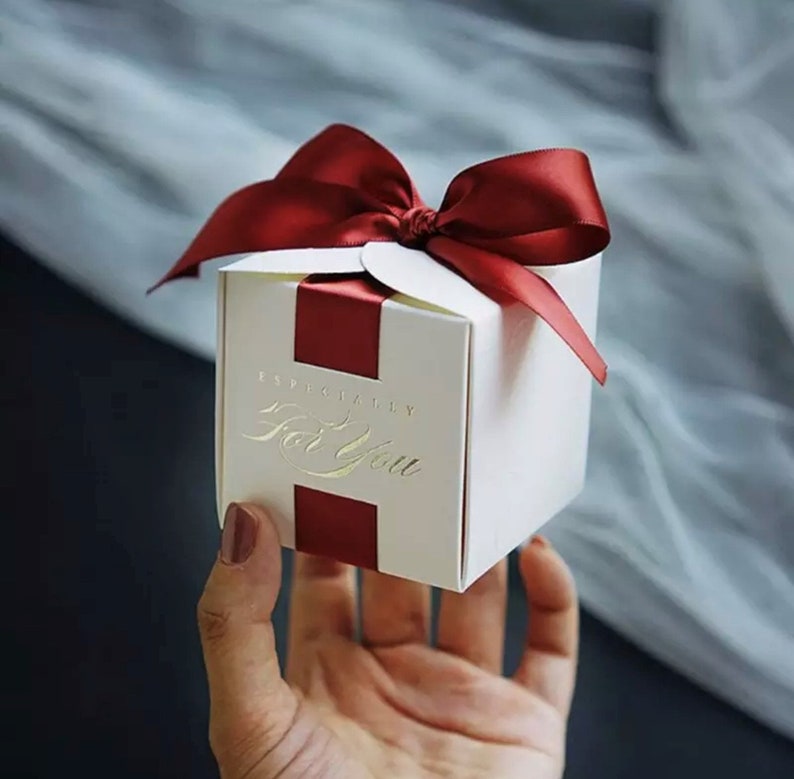 May include: A white gift box with a burgundy ribbon and bow. The box has burgundy stripes and gold lettering that reads "Especially For You". The box is held in a hand.