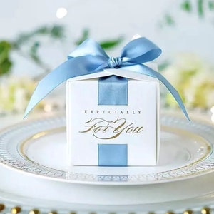 May include: A white gift box with a light blue ribbon and bow, sitting on a white plate with a gold rim. The box has a light blue vertical stripe and the words "Especially For You" in gold script.